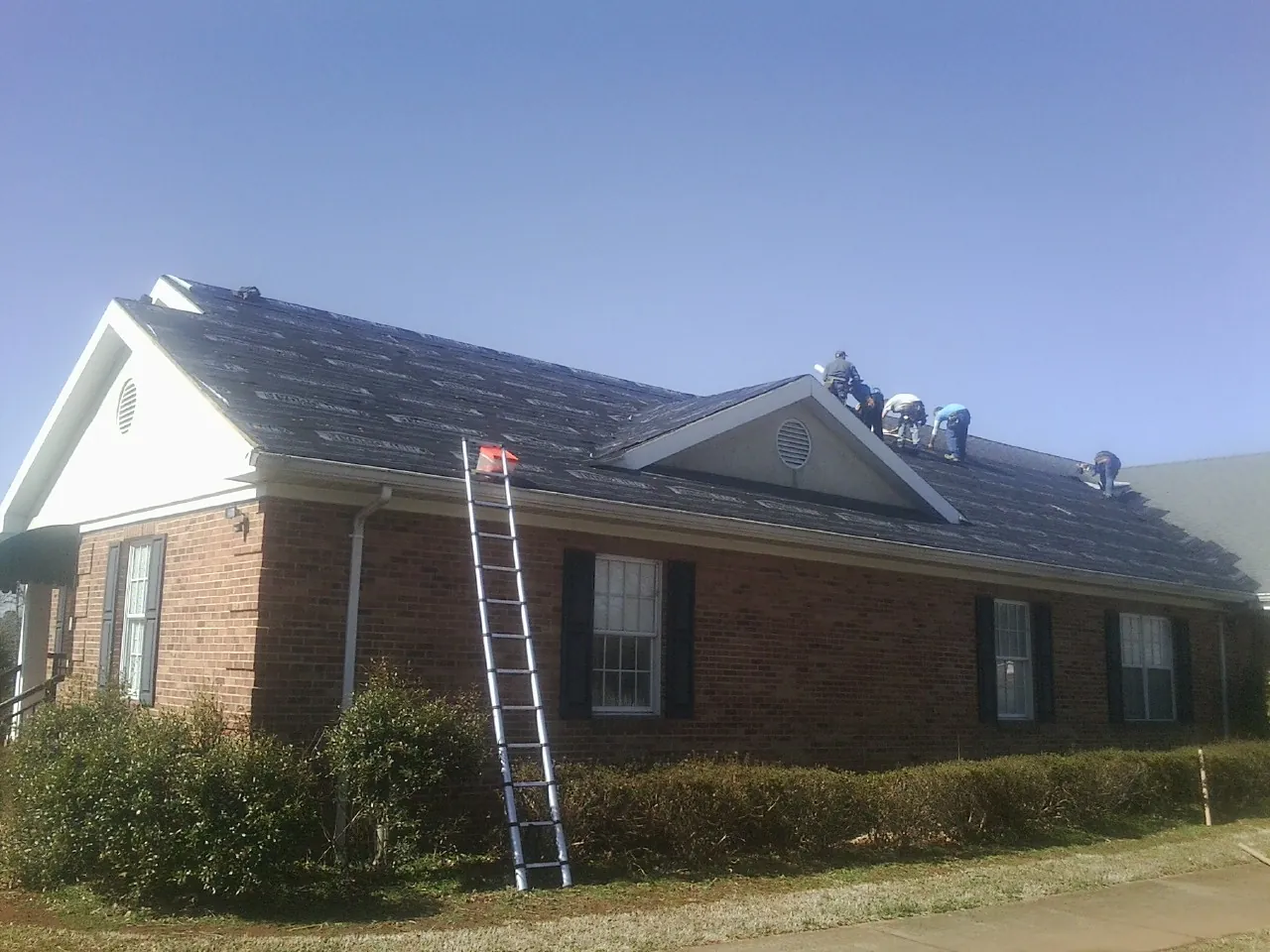 Crew tearing shingles off a brick ranch roof with a ladder set — Emergency Roof Repair work by our crew in Gravity