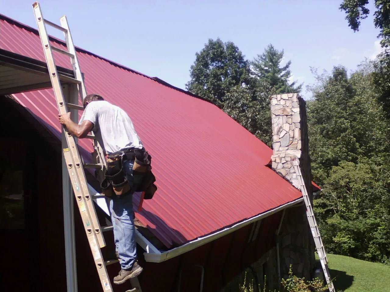 Roofer on a ladder beside a red metal roof and stone chimney — Soffit & Fascia Repair by Roofing Brothers in Gravity